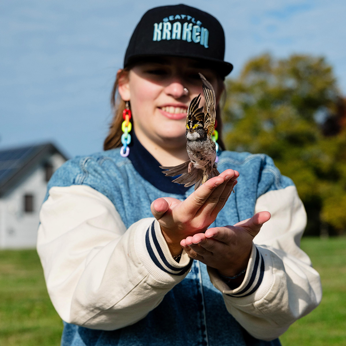 A student in a Kraken hat and bright rainbow earrings holds a bird, smiling