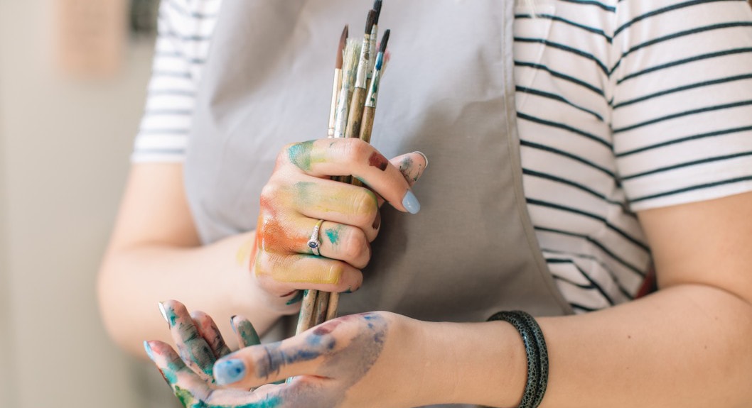Close-up photo of a Chatham University art student wearing an apron and holding paintbrushes in hands that are covered in paint. 