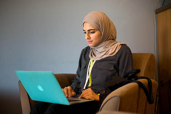 Photo of a young hijabi woman sitting in a chair with a laptop on her lap, working