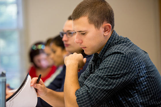 Photo of a male Chatham University student leafing through a pamphlet at a lecture hall desk. 