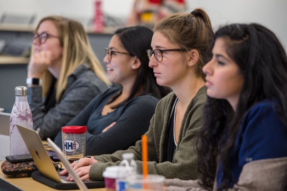 Four female Chatham University students sit at a desk in lecture taking notes and listening. 