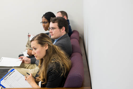 Photo of four counseling psychology students sitting in a row, observing a counseling session off-frame. They are holding clipboards and pens and are taking notes.