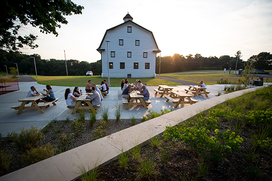 Photo of several people gathering at picnic tables on Chatham University's Eden Hall Campus in front of a white barn. 