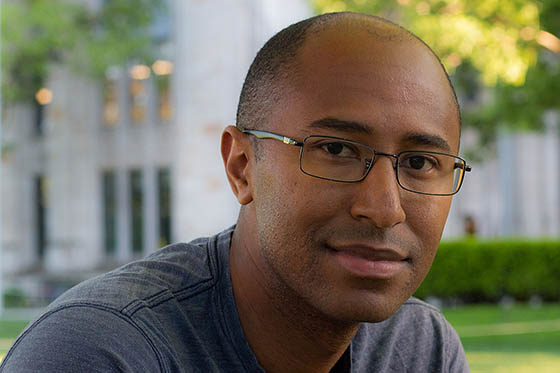 Portrait of Cameron Barnett a Black man with a soft smile, outside in front of greenery