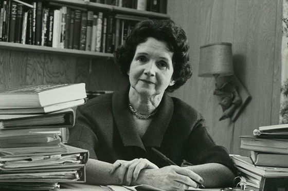 Black and white photo of Rachel Carson sitting behind a desk full of books with pen in hand. 