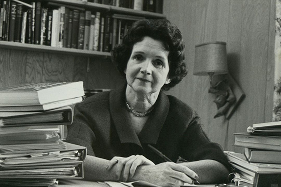 Black and white photo of Rachel Carson sitting behind a desk full of books with pen in hand.