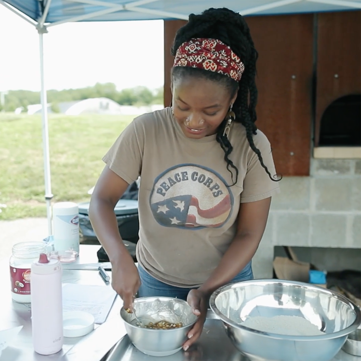 Photo of a young Black woman wearing a Peace Corps shirt, smiling and stirring food in a bowl at the Eden Hall Campus Bread Oven