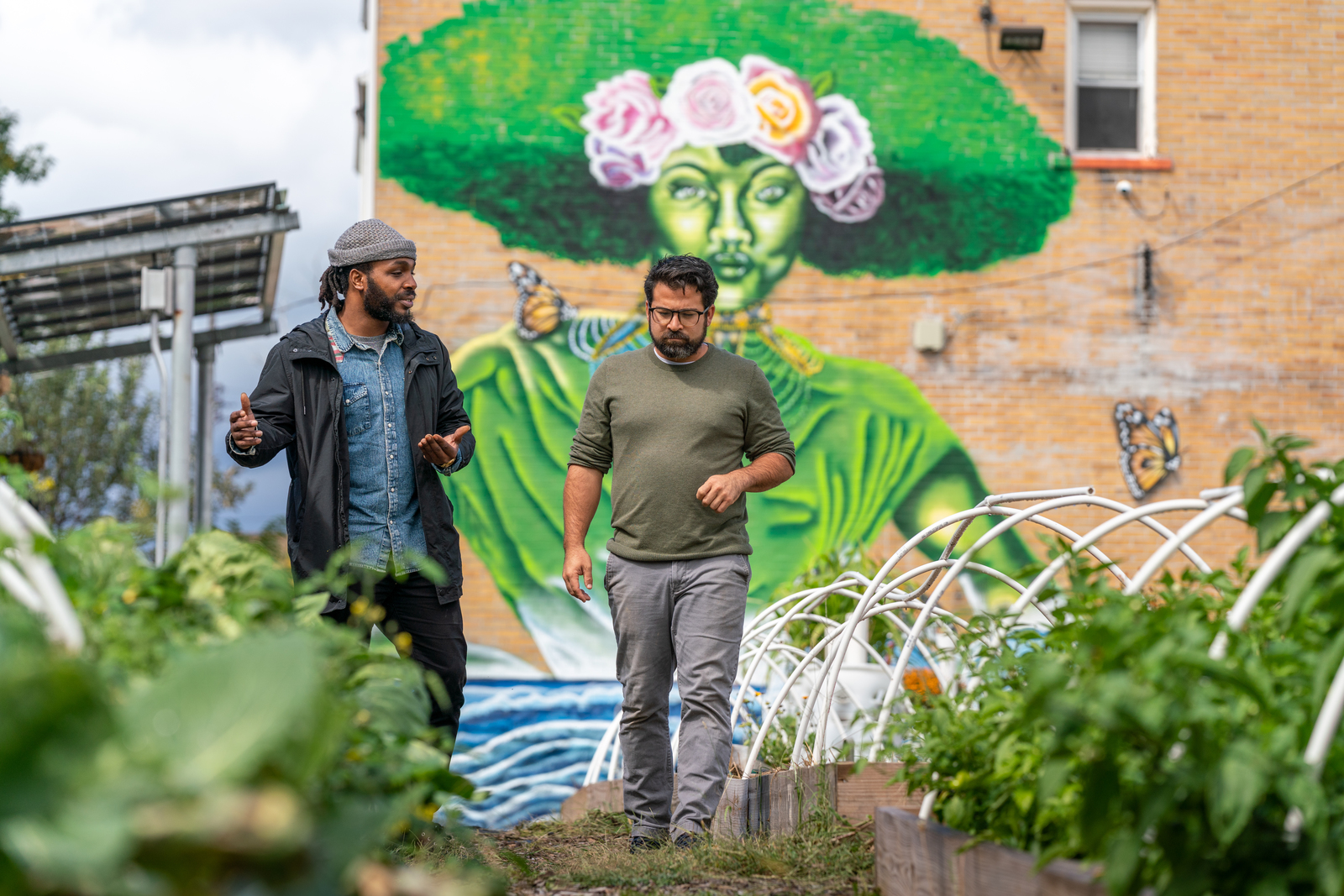 Photo of two men walking through a city garden, with a large colorful mural on the brick wall behind them