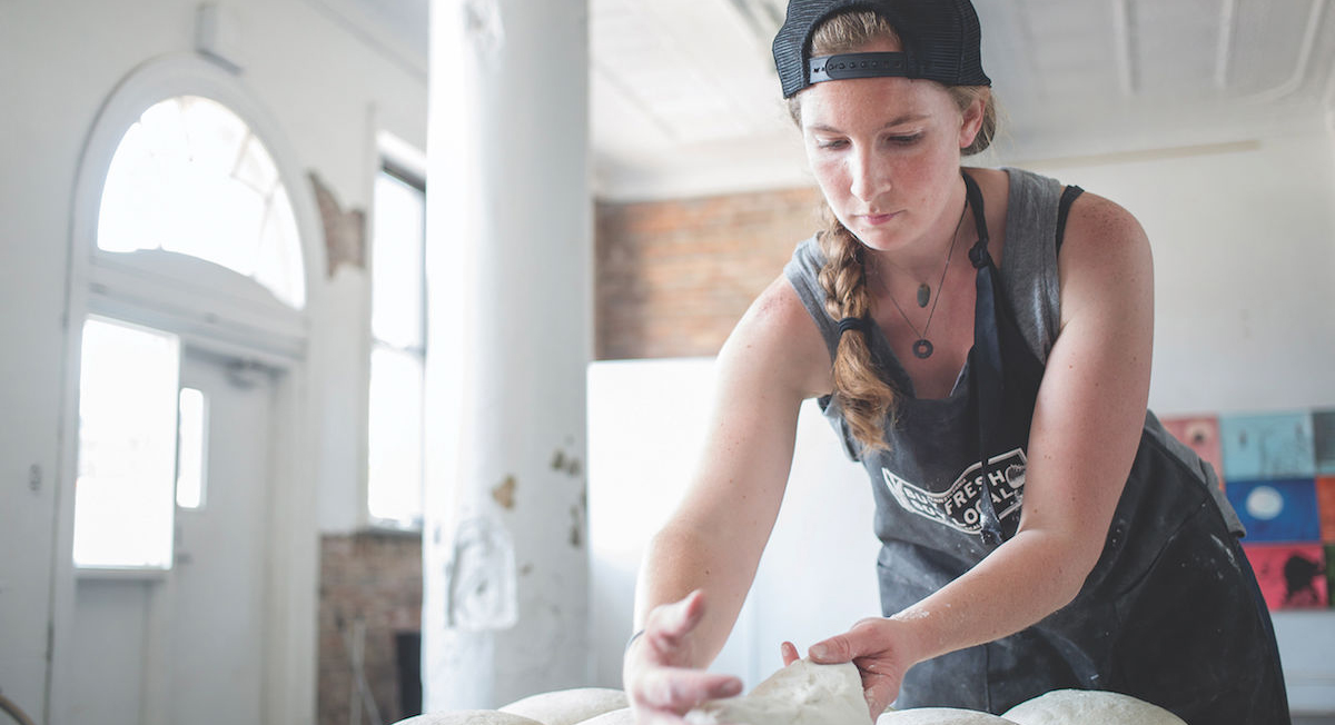 Woman wearing backwards hat and apron arranges bread dough on a floured table. 