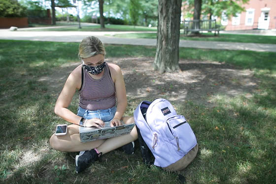 Photo of a masked student sitting on the ground at Shadyside Campus, working on her laptop