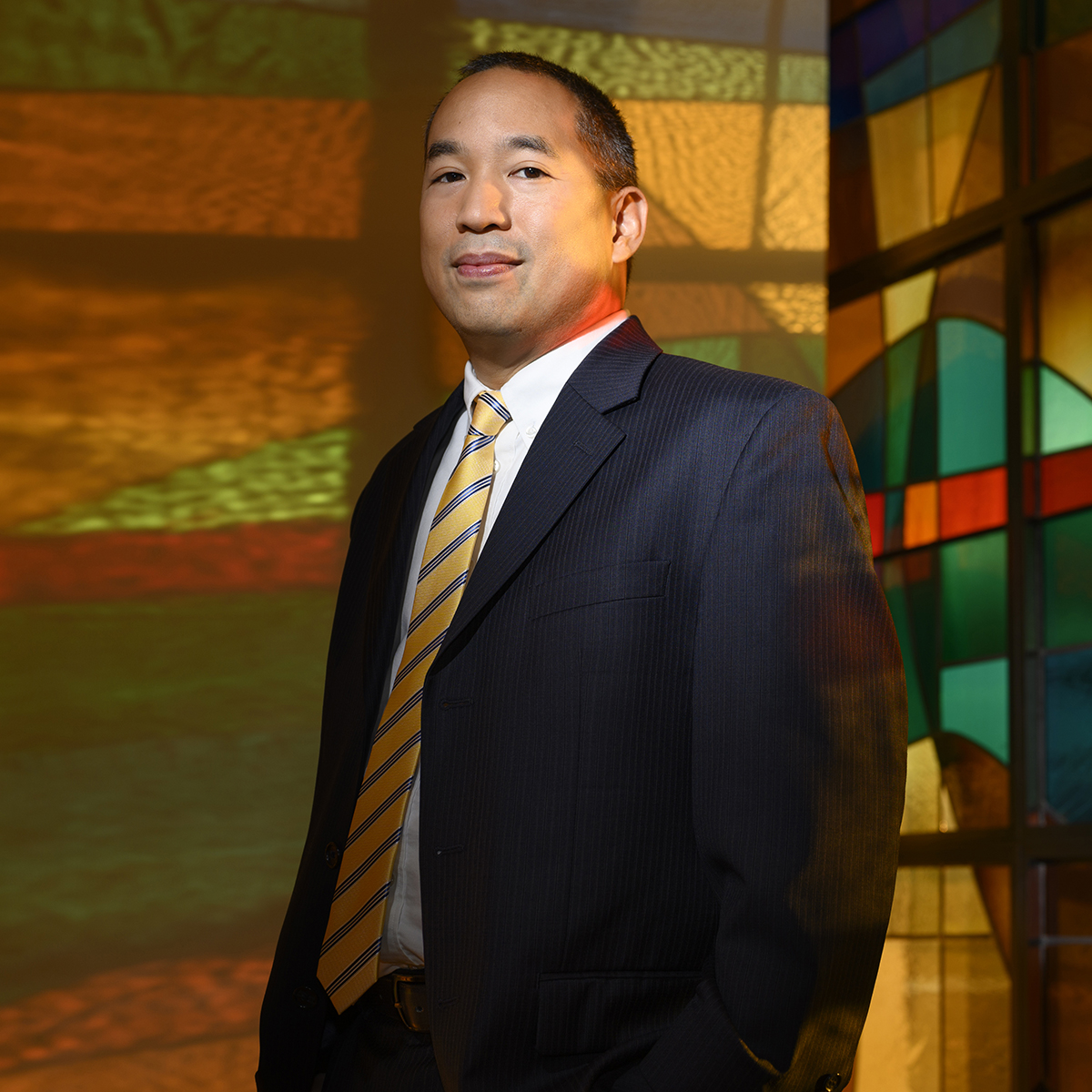 Photo of a smiling man in a suit, standing in front of colorful stained glass