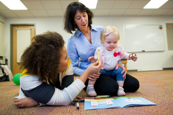 Photo of an instructor and female student on the floor, interacting with a small child looking at a picture book