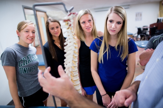Photo of an Instructor pointing at a spinal skeletal model in the classroom, with Chatham University circling around him. 