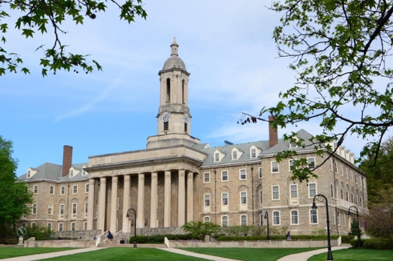 Photo of "Old Main," a grey stone academic hall with a bell tower, at Penn State framed by tree branches with green leaves.