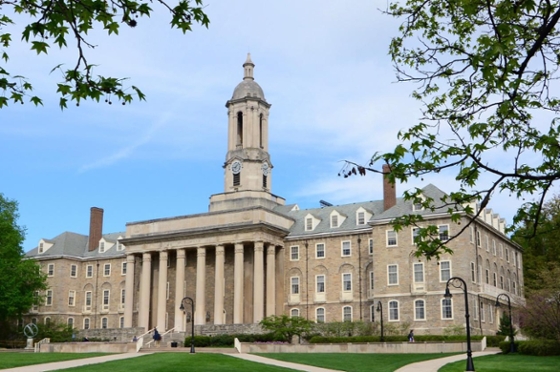 Photo of "Old Main," a grey stone academic hall with a bell tower, at Penn State framed by tree branches with green leaves. 