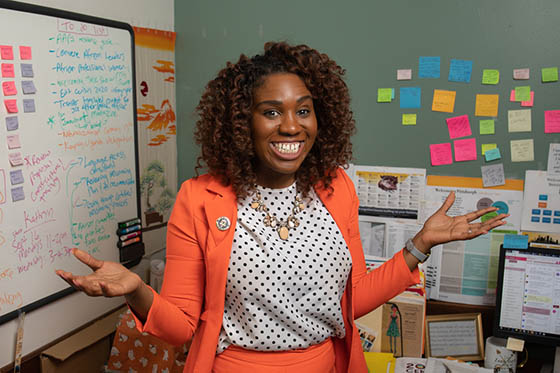 Photo of a Chatham University MSUS+MBA graduate in a professional suit, posing in front of her desk with a white board and post-it notes in the background