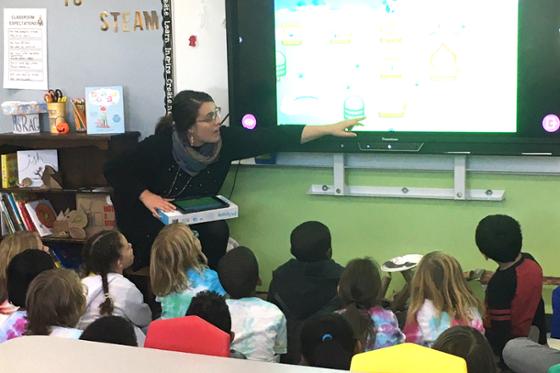 Photo of an elementary school teacher speaking to group of children sitting in front of Smart Board in classroom.