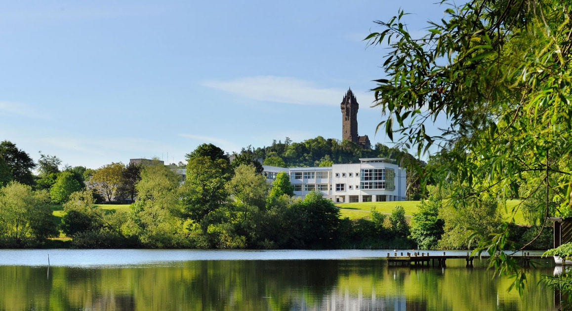 Photo of green forests and a calm lake, with a white building and a tall tower behind it: The University of Stirling in Stirling, Scotland.