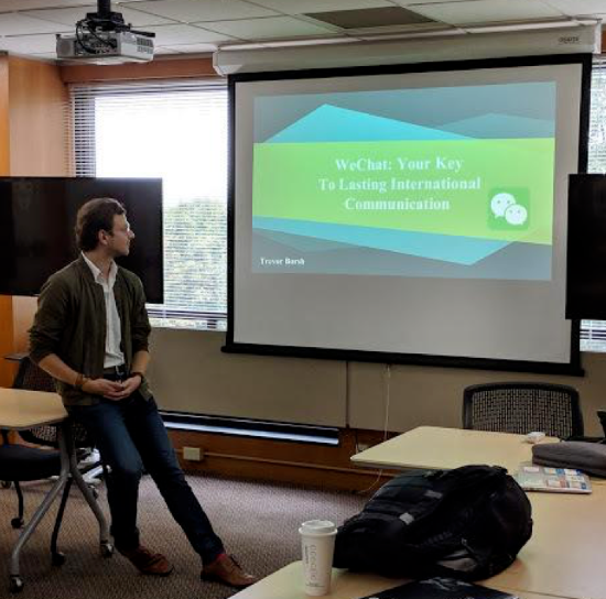 Photo of a PowerPoint title slide (WeChat: Your Key to Lasting International Communication) displayed on a screen in a classroom, with a man leaning against a desk in front of it