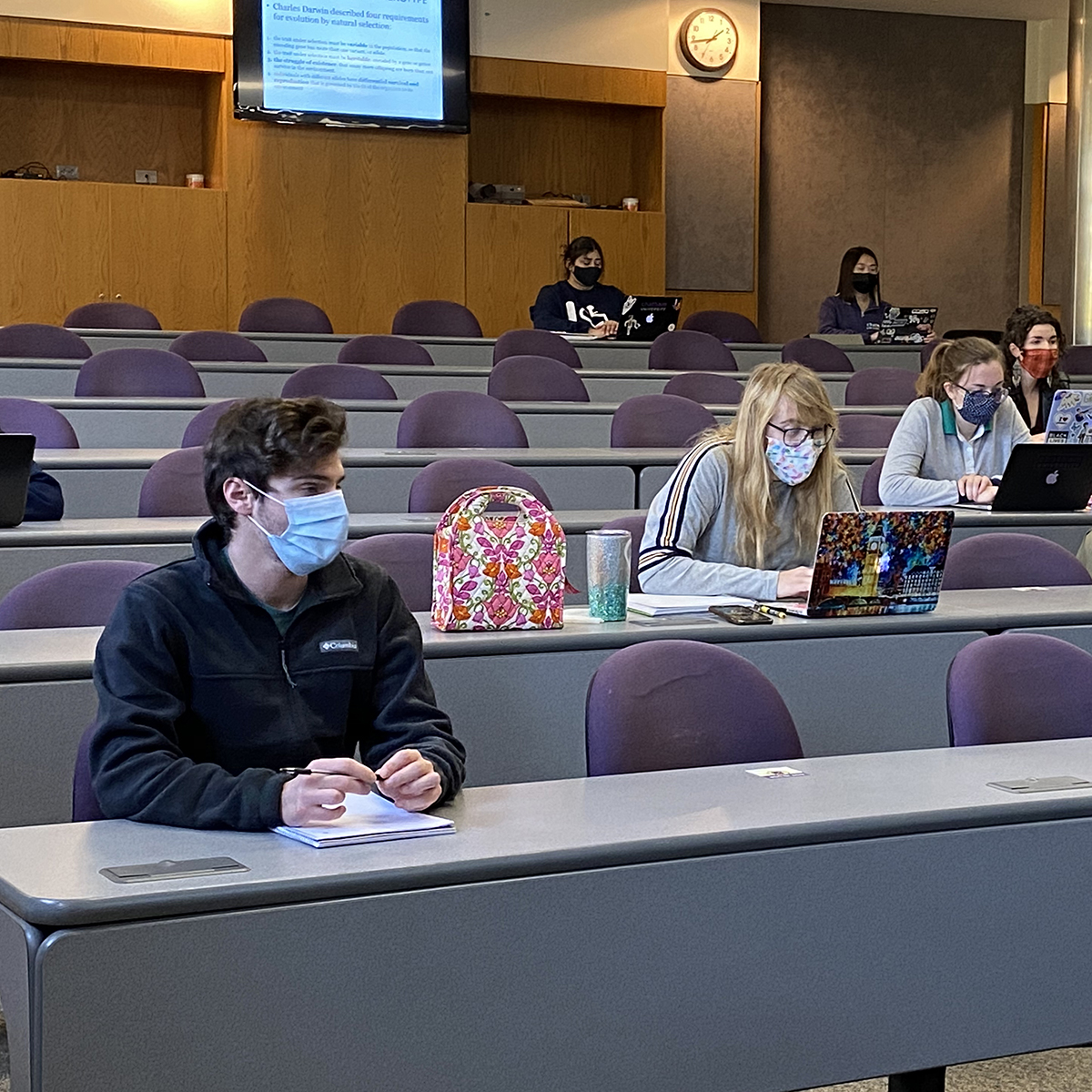 Photo of masked students seated at a distance from one another in a lecture hall, working in notebooks and on laptops