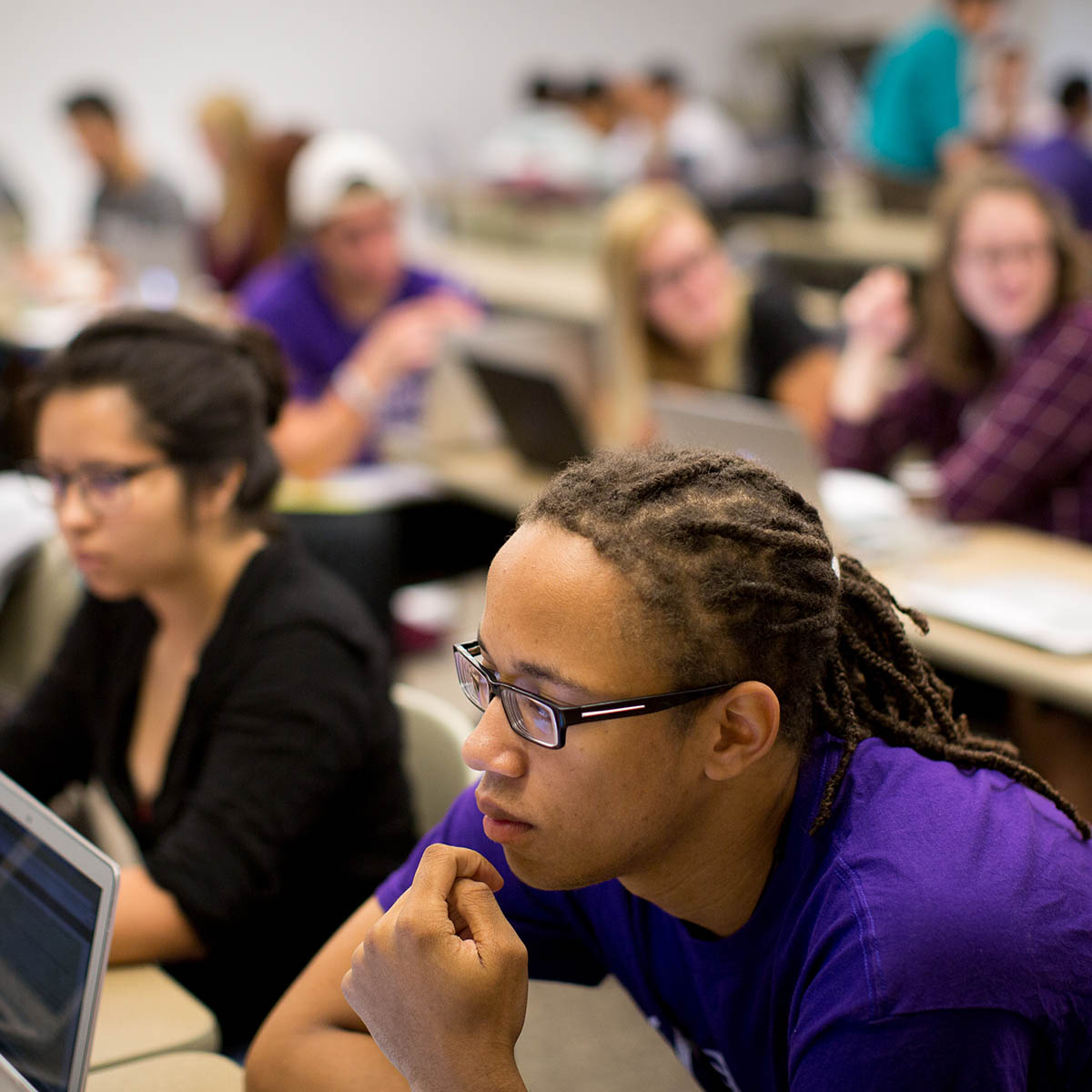 Photo of students listening in a classroom, with a young Black man with dreads, glasses, and a purple Chatham shirt in the foreground