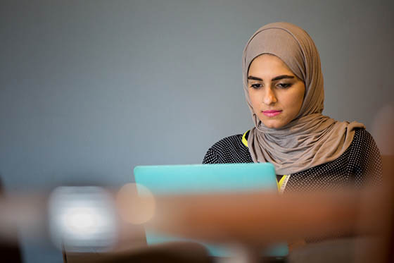 Photo of a Chatham University student wearing a hijab, smiling softly while working on her laptop in Cafe Rachel