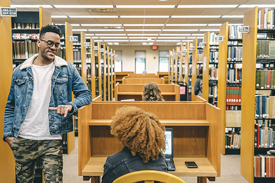 Photo of two students chatting at a cubicle in Jennie Mellon King library, between the stacks of library books.