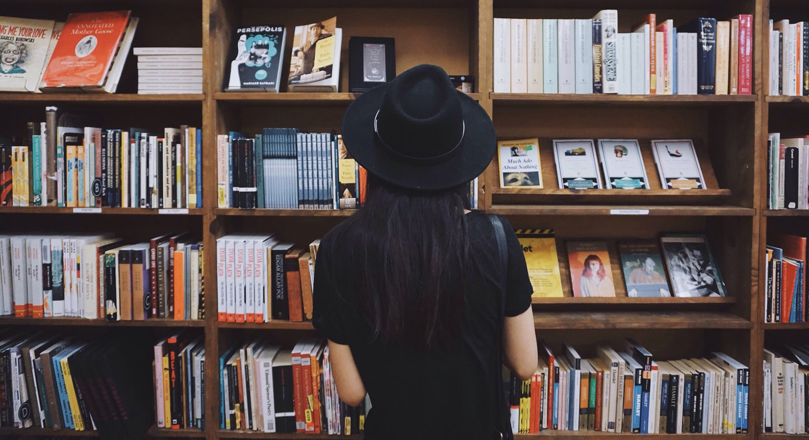 Photo of a woman wearing a black wide-brimmed hat and black t-shirt stands in front of a colorful bookshelf. 