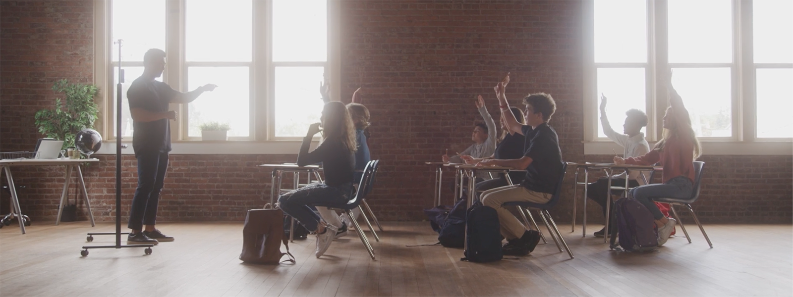 Photo of a teacher at the front of a classroom, with students raising their hands