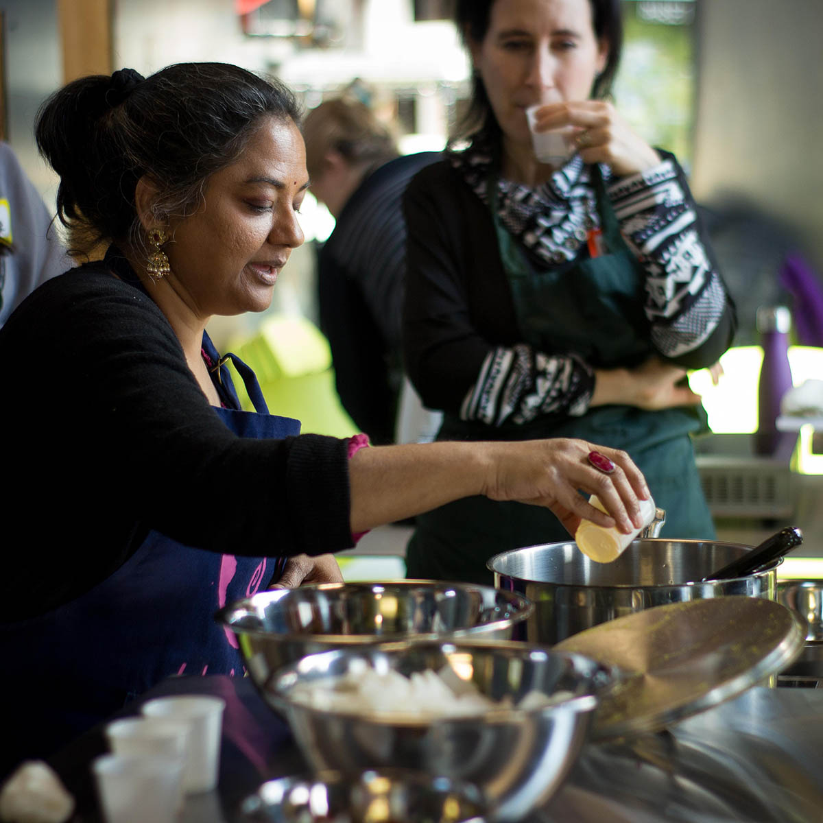 Photo of a South Asian woman demonstrating how to cook, while a class observes