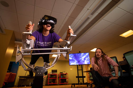 Photo of a student wearing a purple Chatham University shirt, in a virtual reality machine, while a professor watches on