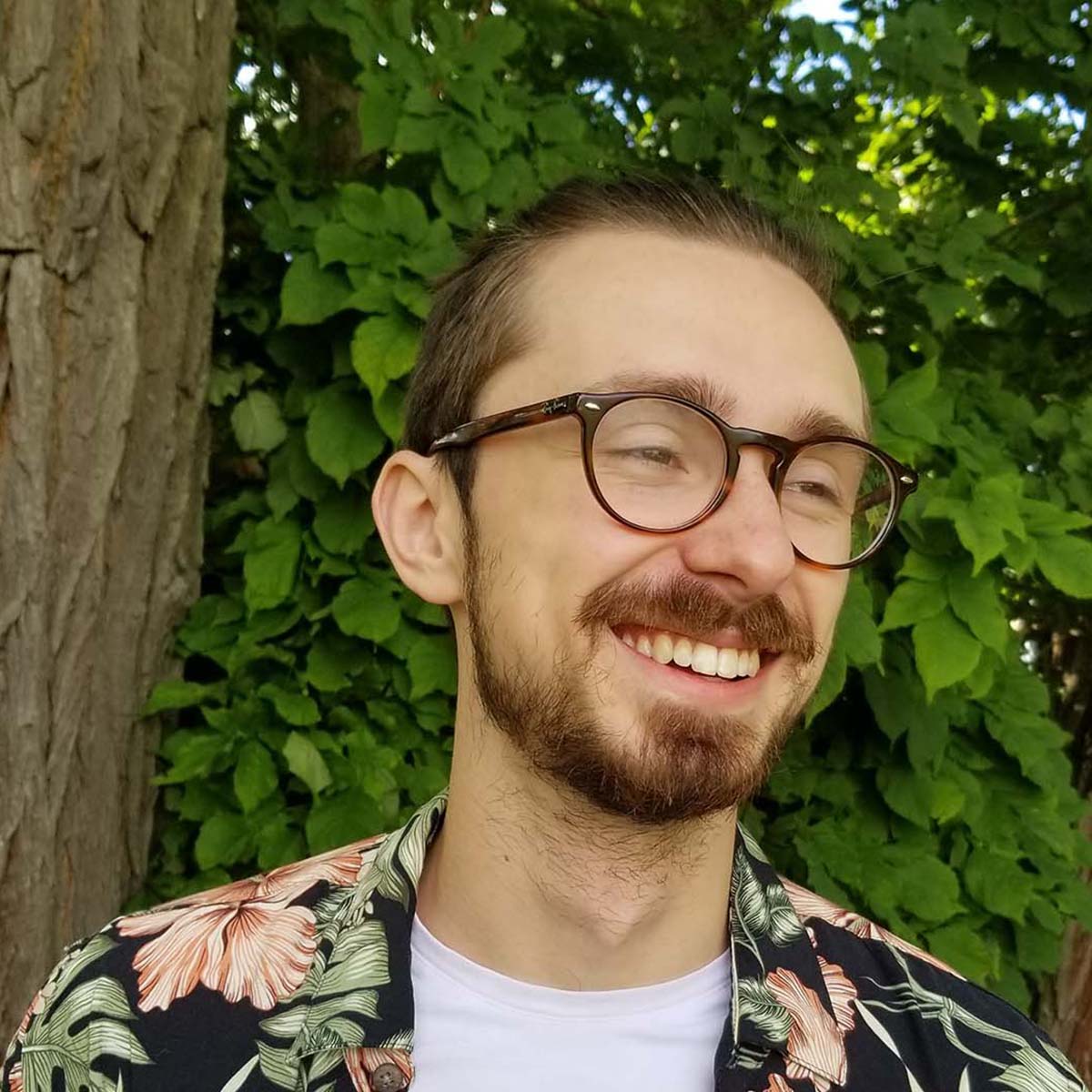 Photo of Carson Custer, a young white man with glasses and a bright floral shirt, smiling in front of greenery