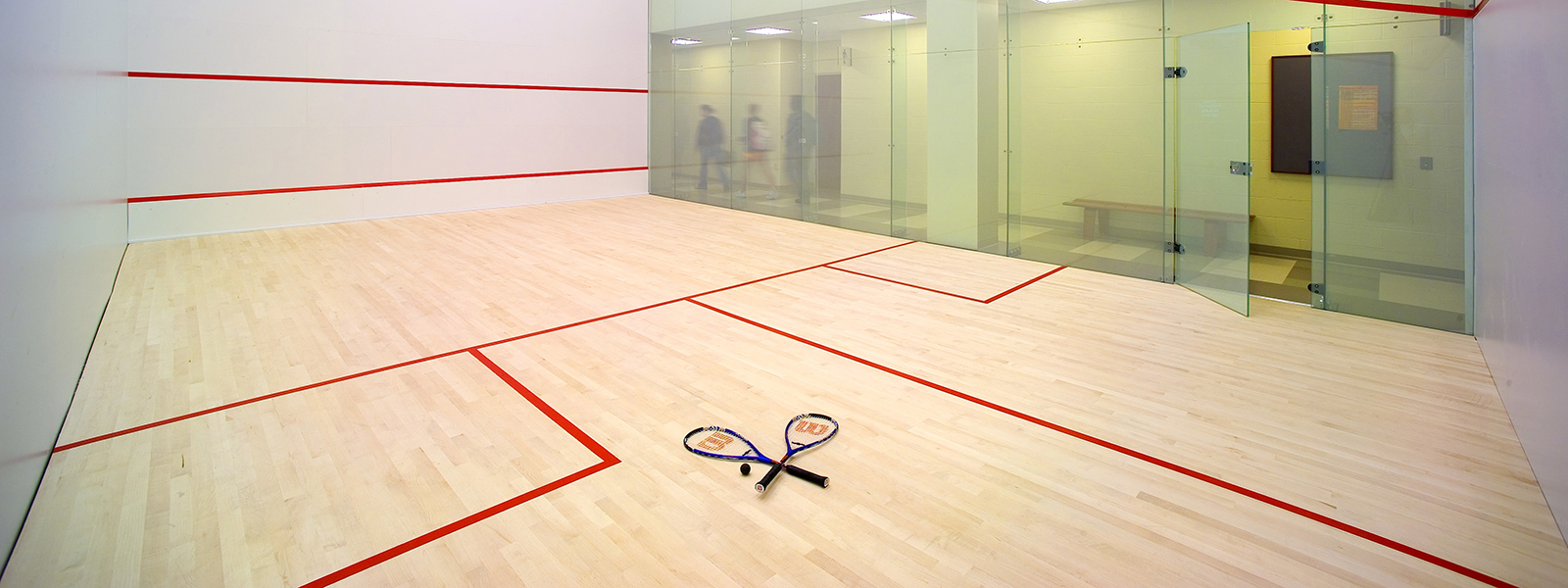 Photo of a squash court in the gym, with a ball and two rackets on the ground. 