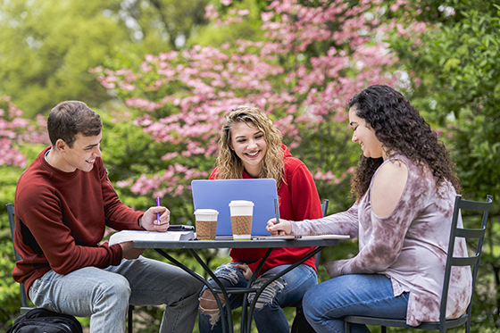 Photo of three Chatham University students sitting at a patio table outside working together on laptops and smiling. 