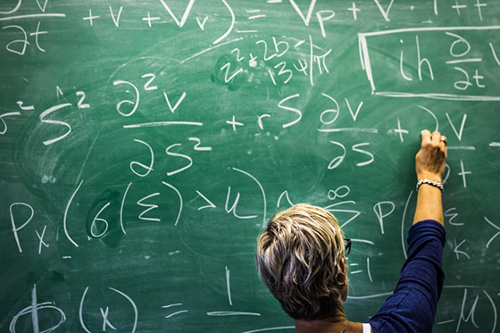 Photo of a blonde short-haired woman writing mathematic equations on a green chalkboard. 