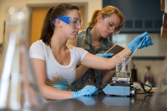 Photo of two female students in protective lab gear performing chemistry experiments