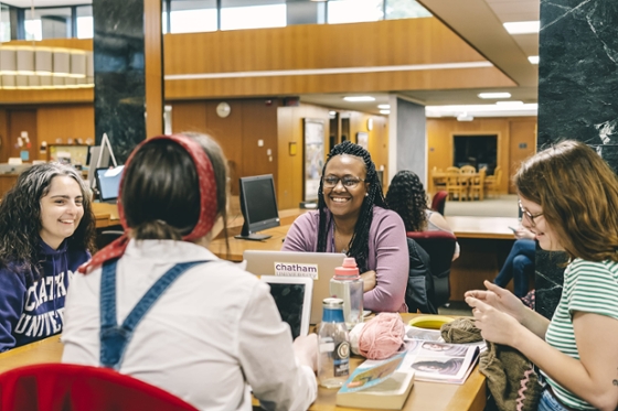 Photo of four students sitting around a table in the library, talking and laughing, with laptops and books and knitting on the table