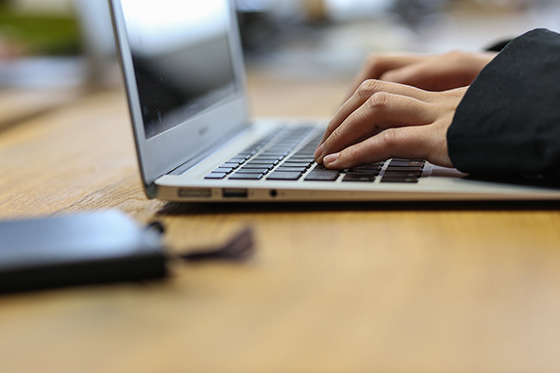 Close-up of a photo typing on a laptop, with a notebook on the table