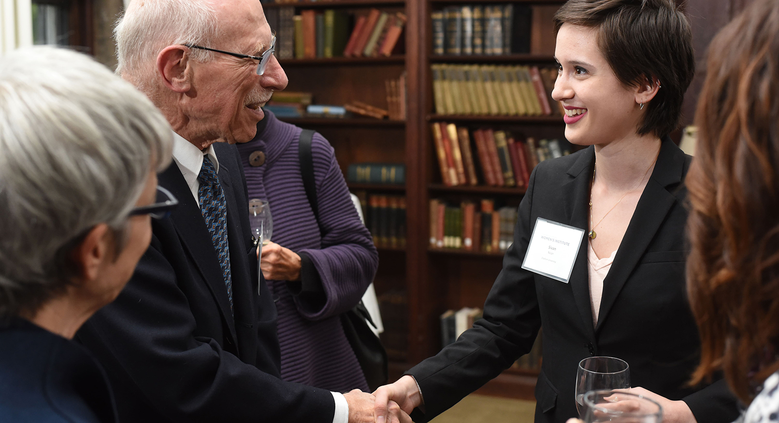Photo of a Chatham University student in a black blazer and name tag shaking the hand of an elderly man at a networking event. 