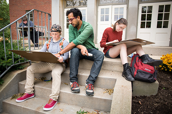 Photo of a Chatham University art professor sitting on steps outside with students who are drawing on art boards. 