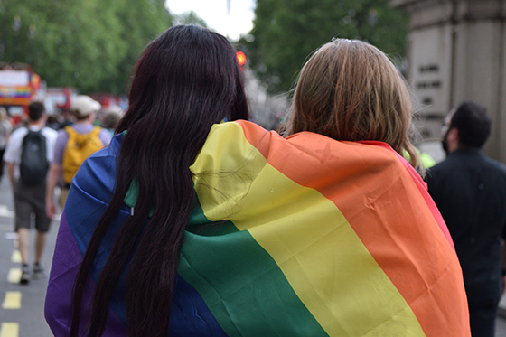 Photo of two people standing under a rainbow LGBTQIA+ flag at a parade