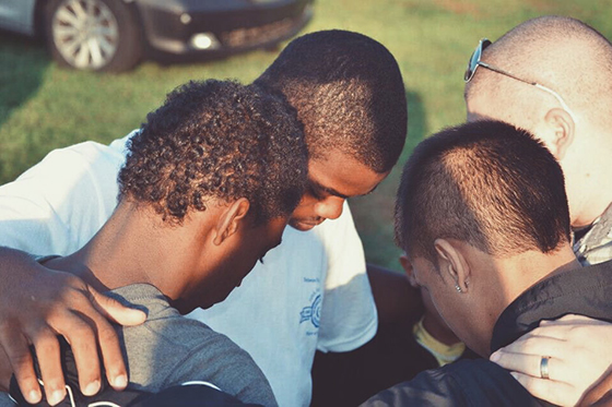 Photo of four young Black boys bowing their heads together in prayer outside