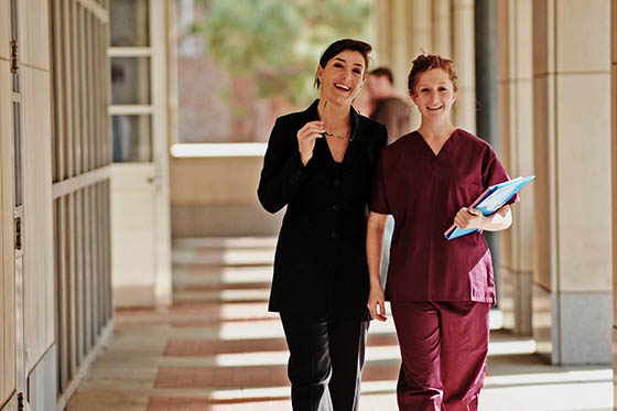 Photo of a nurse in maroon scrubs walking with a woman in a black suit in an outdoor hallway