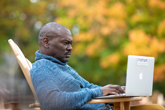 Photo of a Chatham University male student working on his laptop outside, with autumnal leaves in the background.