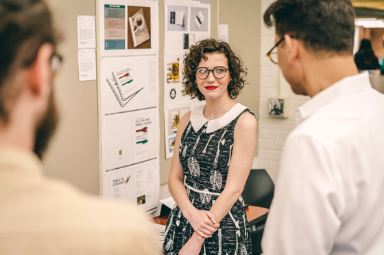 Photo of a female Chatham University student standing in front of her presentation, as two professors offer thoughts.