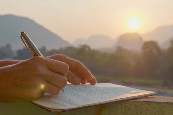 Photo of a person's hands, as they write in a notebook with a sunset in the background