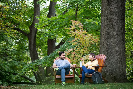 Photo of two Chatham University students sitting outside drinking coffee on Adirondack chairs under tall oak trees on green grass. 