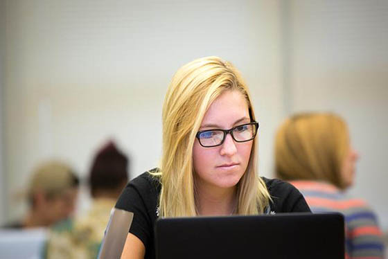 Photo of a white blonde woman in glasses working on a computer in a classroom