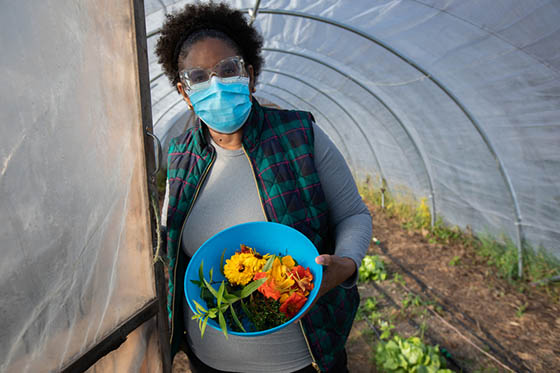 Photo of a Chatham University student in a mask, holding a bowl of harvested produce at the door of a greenhouse
