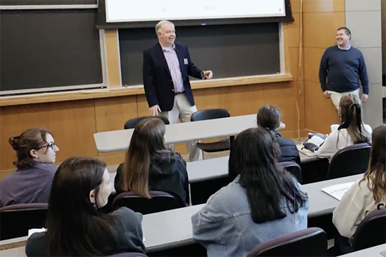 A professor speaks to a group of students seated in a lecture hall, while another professor stands nearby smiling